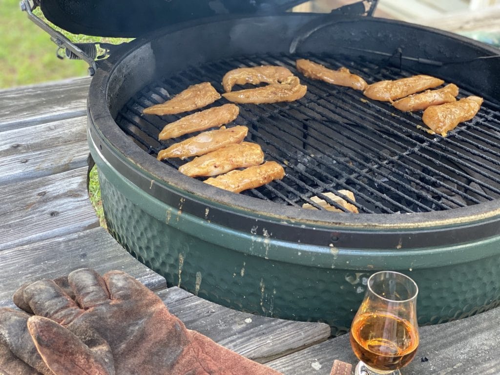 chicken tenders on a big green egg with a glass of whiskey on a rough wood table with a glass of whiskey and leather gloves