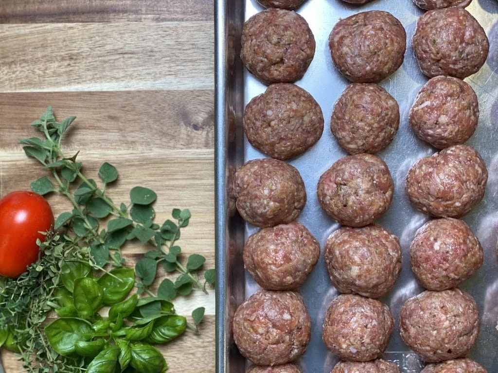 Raw meatballs on cookies sheet with herbs beside them.
