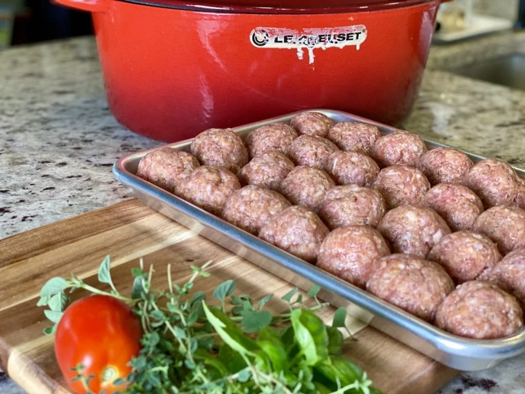 raw meatballs with herbs in the foreground and a le creuset Dutch oven in the background.