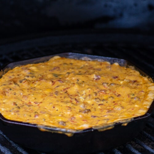 A skillet with melted cheese dip on the grill. The cheese dip is orange and slightly spilling over the edge. It is elected with bits of tomato and sausage.