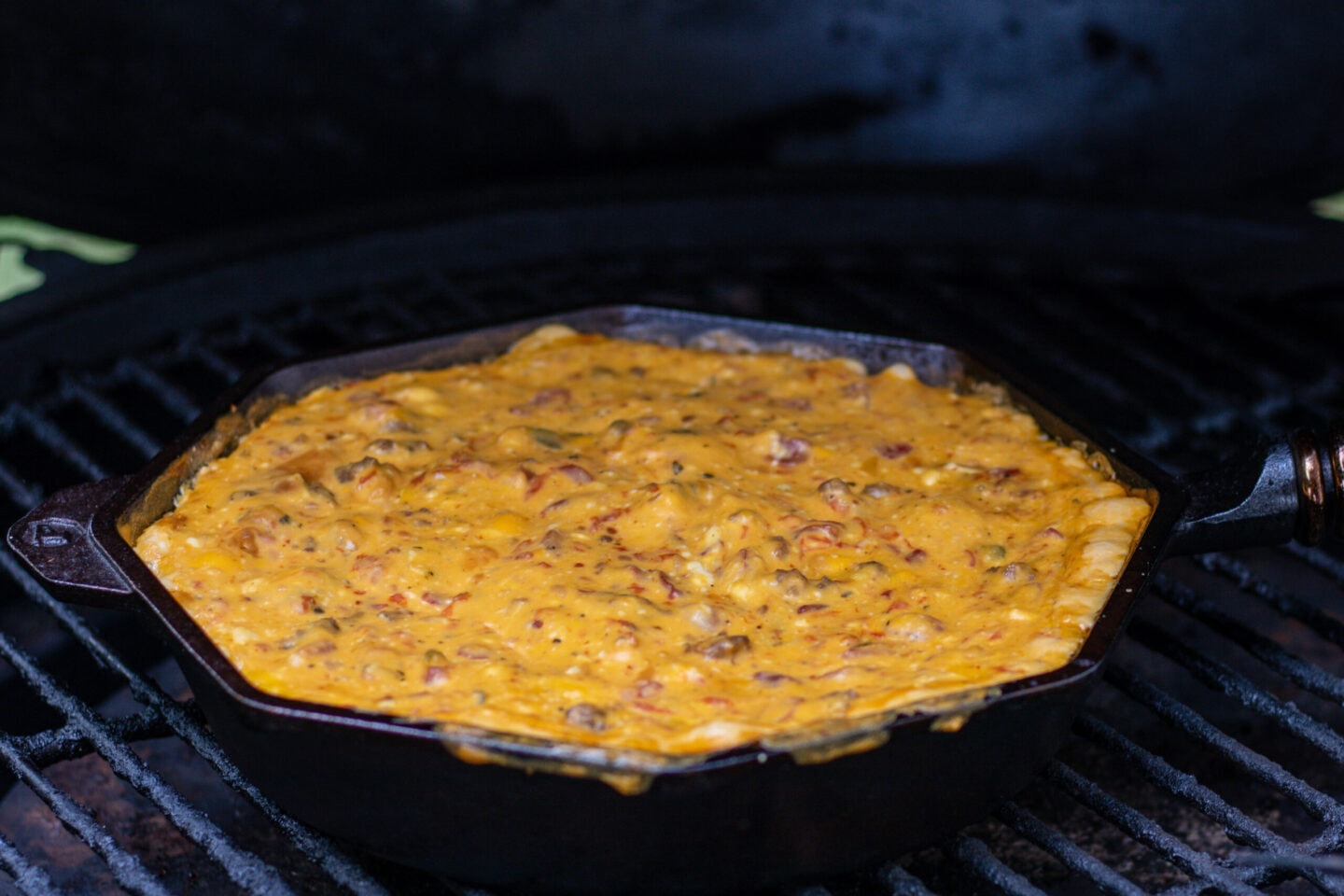 A skillet with melted cheese dip on the grill. The cheese dip is orange and slightly spilling over the edge. It is elected with bits of tomato and sausage.