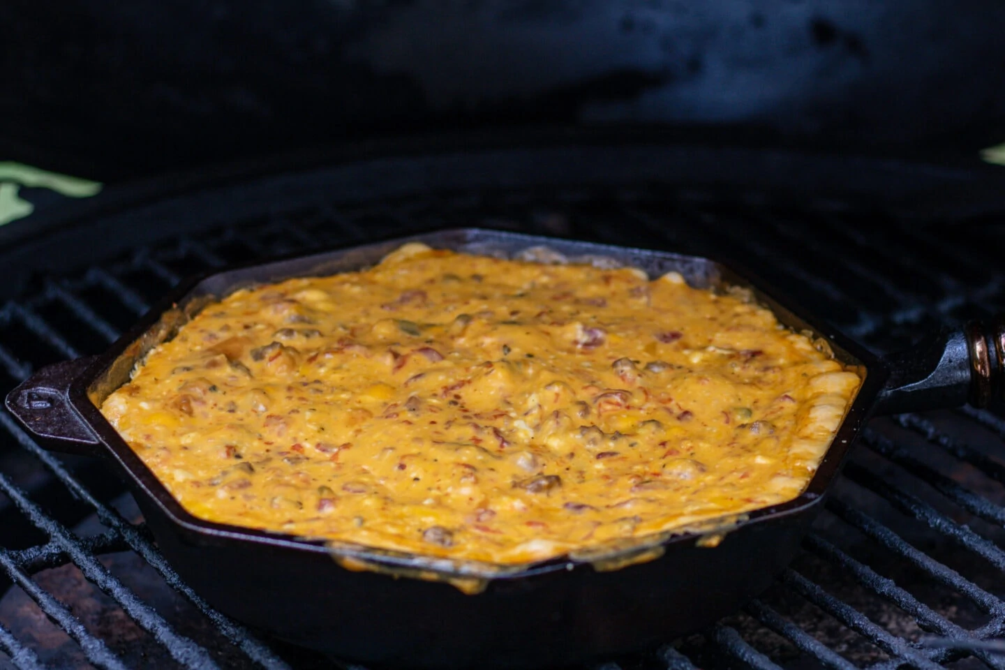 A skillet with melted cheese dip on the grill. The cheese dip is orange and slightly spilling over the edge. It is elected with bits of tomato and sausage.
