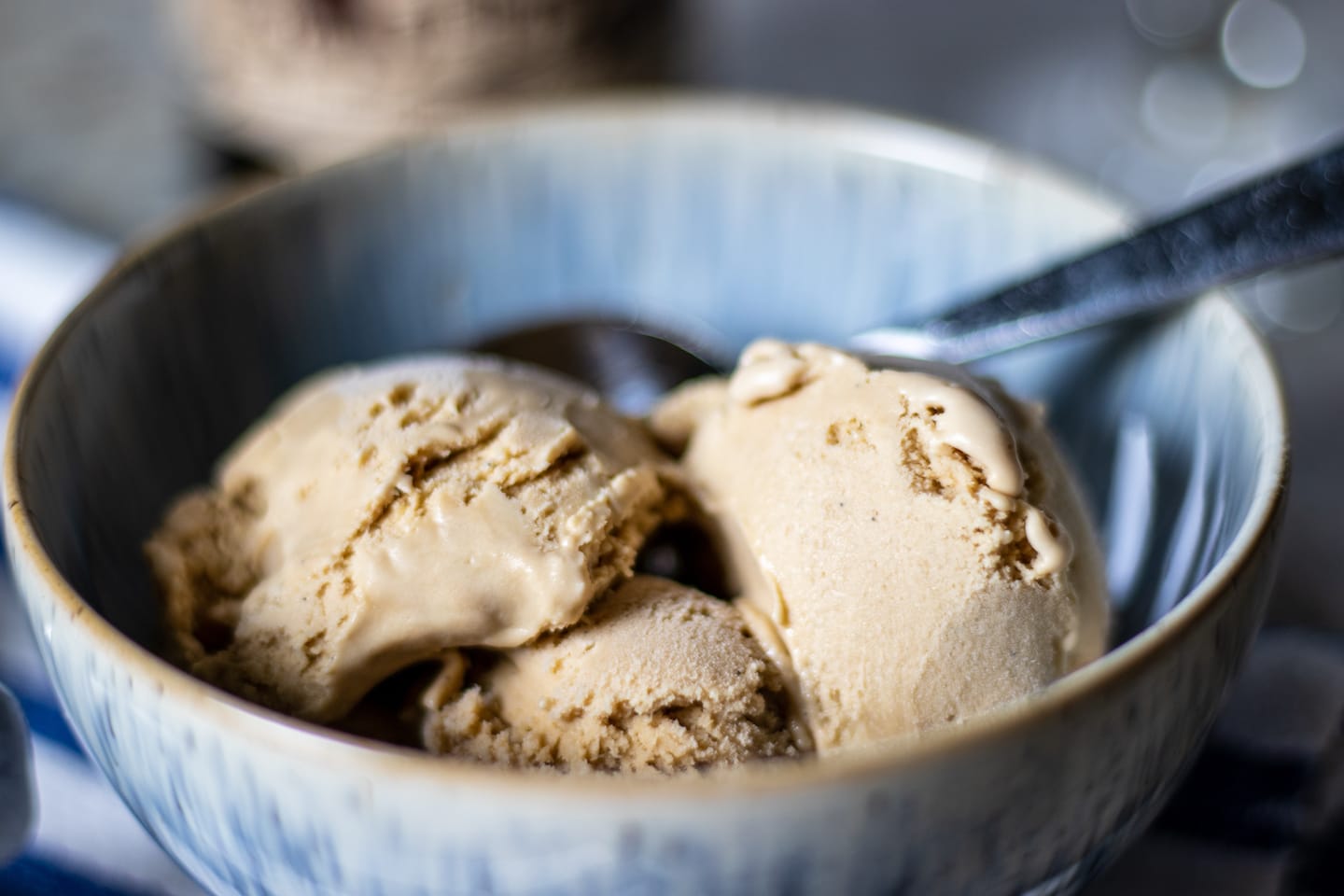 A blue specked bowl with three scoops of tan ice cream and a metal spoon to sticking out of the top right ready to be used.