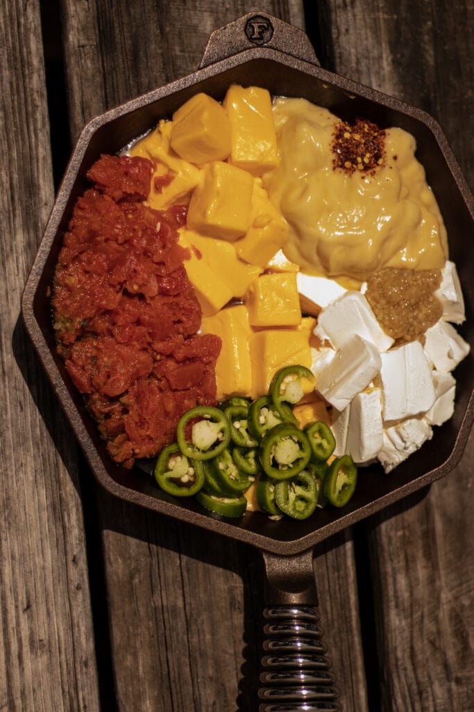 A skillet with tomatoes, cream cheese, velveeta, jalapeños, cream of chicken soup, garlic, and red pepper flakes on a weathered wooden table.