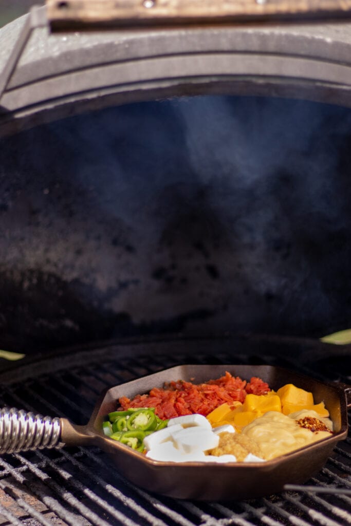 A skillet with tomatoes, cream cheese, velveeta, jalapeños, cream of chicken soup, garlic, and red pepper flakes on a grill with a small trail of smoke