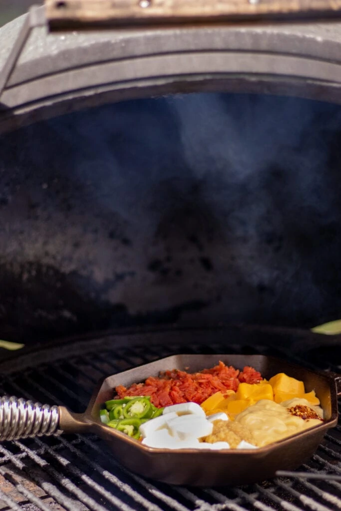 A skillet with tomatoes, cream cheese, velveeta, jalapeños, cream of chicken soup, garlic, and red pepper flakes on a grill with a small trail of smoke