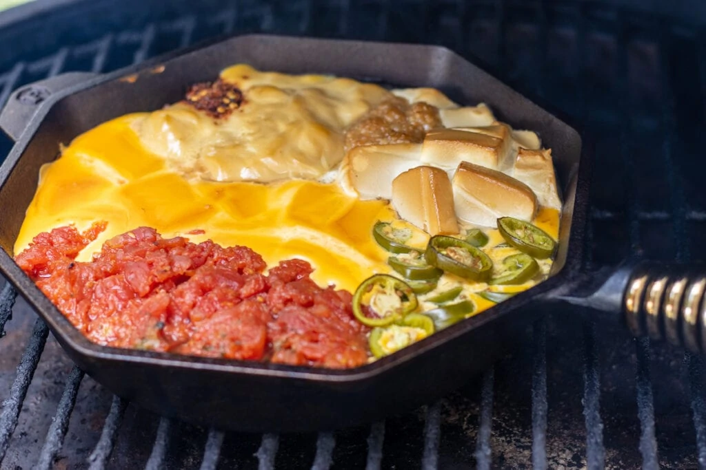 A skillet with tomatoes, cream cheese, velveeta, jalapeños, cream of chicken soup, garlic, and red pepper flakes on a grill smoked a slightly melted.
