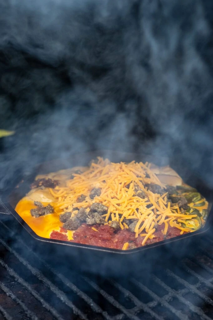 A skillet with tomatoes, cream cheese, velveeta, jalapeños, cream of chicken soup, garlic, and red pepper flakes on a grill topped with shredded cheddar and crumbled sausage surrounded by smoke