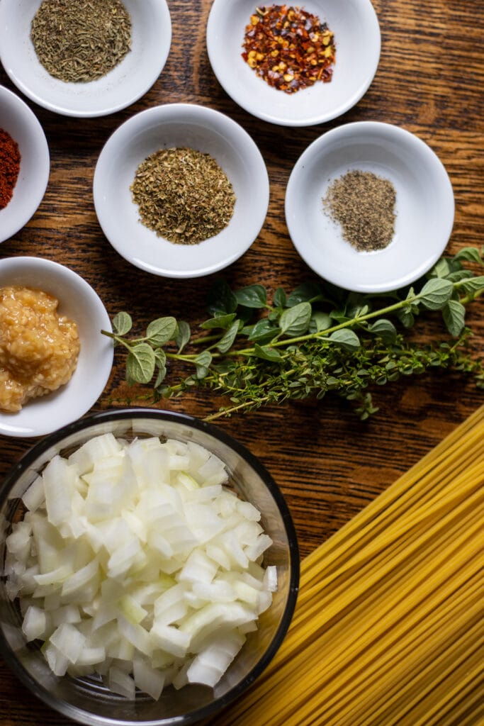 Dried spices, herbs, and garlic in small white bowls, a larger bowl of diced onion is on the bottom left, dried spaghetti is on the bottom right and a bundle of fresh thyme and oregano are in the middle.