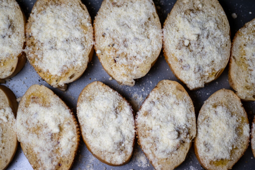 Slices of French bread on a cookie sheet topped with parmesan cheese on top of butter and herbs