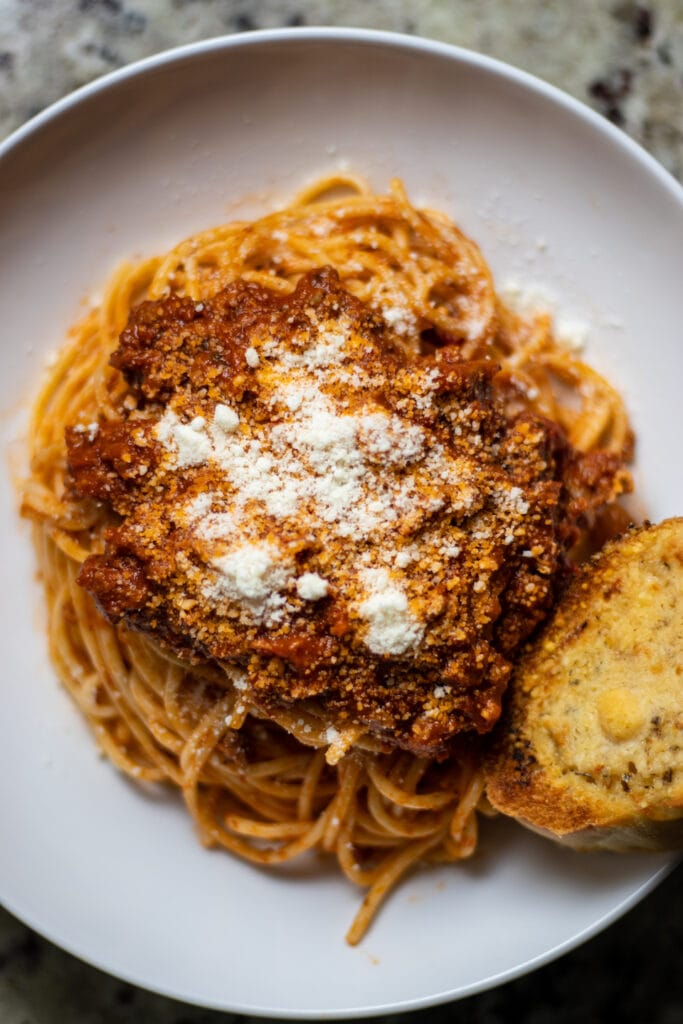 Spaghetti topped with a red marinara sprinkled with grated Parmesan cheese. A piece of garlic bread is on the bottom right.