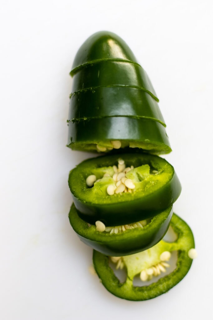 A sliced green jalapeño on a white cutting board. The pepper is sliced across and is vertical on the board so the bottom end of the jalapeño is pointed to toward the top of the frame. It is bright green and the seed and membrane are visible and fresh looking.