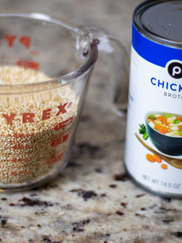 A pyrex measuring cup with dried quinoa on the left and a public can of chicken broth half off frame to the right. 