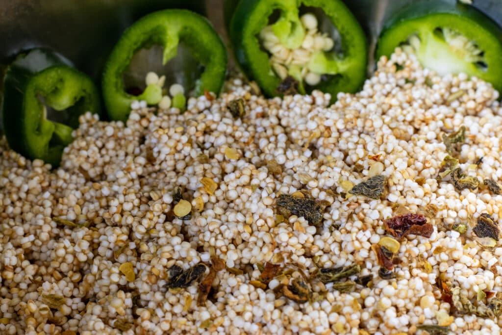 Close up of quinoa in a pot with chicken stock. Dried green peppers are on top and sliced jalapeño peppers line the pot int he background.