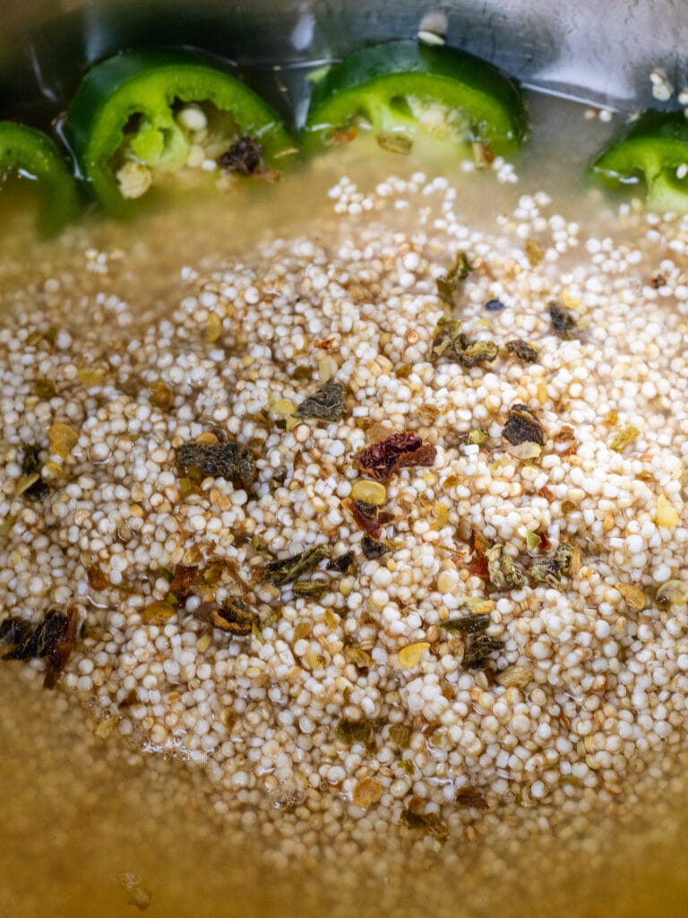 Close up of quinoa in a pot with chicken stock. Dried green peppers are on top and sliced jalapeño peppers line the pot int he background.