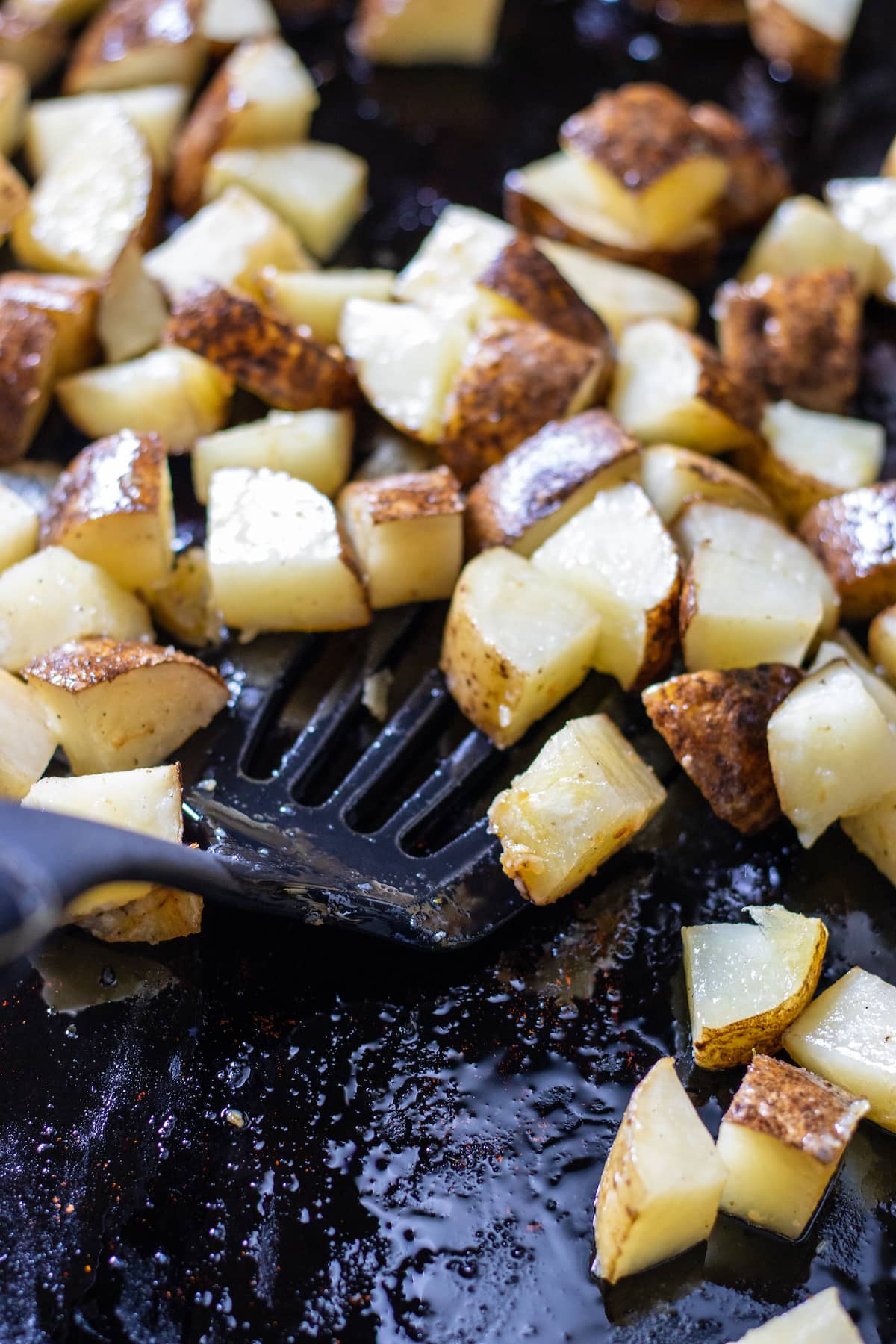 partially cooked diced potatoes being flipped with a black spatula on a cookie sheet