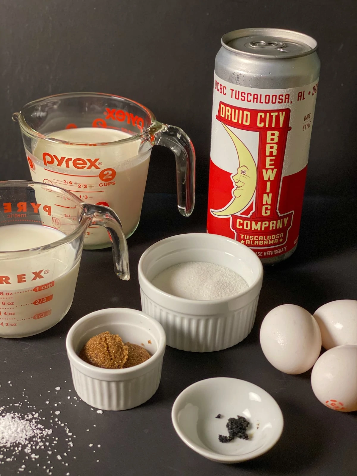 cream, milk, a can of beer with a cresent moon, sugar, brown sugar, scraped vanilla bean, and eggs on a black background.