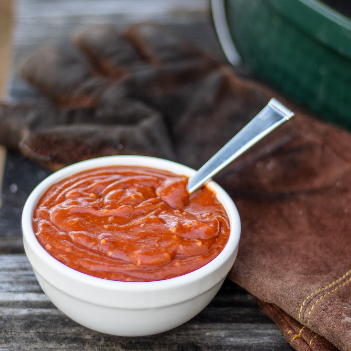 a bowl of BBQ sauce outside on a wooden table by some leather grilling gloves