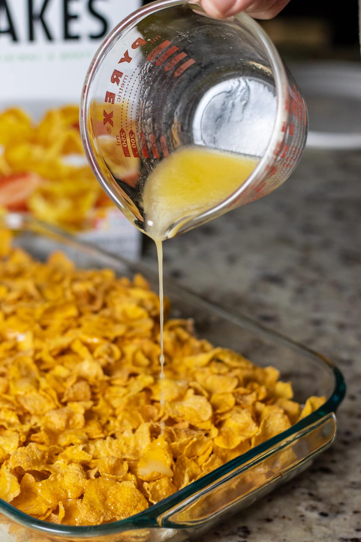 Butter being drizzled from a glass measuring cup over the corn flakes topping the hashbrown casserole.