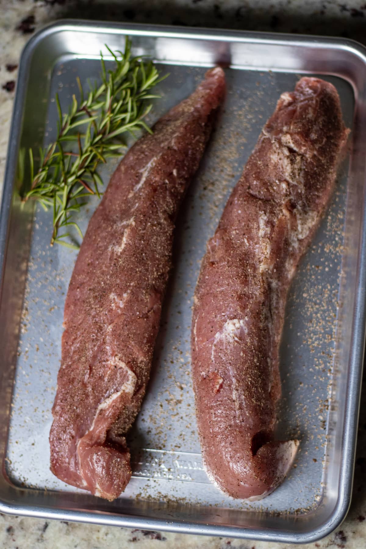 raw pork tenderloin rubbed with salt and pepper an on a small aluminum cookie sheet with some rosemary